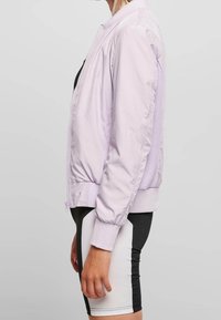 Jasnolawendowa bomberka o gładkim wykończeniu, z żebrowanymi mankietami i siateczkowymi detalami po bokach. Widoczne zapięcie na zamek i elastyczny dół.