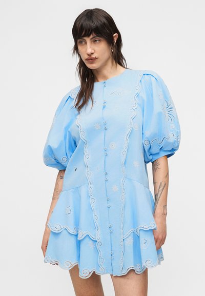 Γυναίκα με σκουρόχρωμα μαλλιά και τατουάζ που φοράει ένα ανοιχτού γαλάζιου χρώματος φόρεμα με κουμπιά, φουσκωτά μανίκια και κεντήματα με μοτίβα ήλιου, φεγγαριού και σύννεφων.