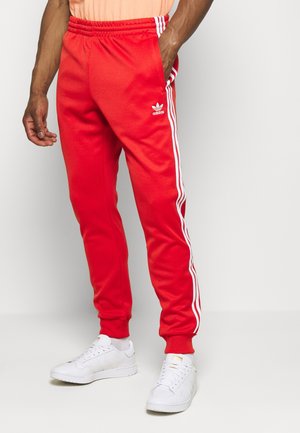 Mann trägt rote Adidas-Trainingshose mit weißen Streifen an den Seiten und weißen Sneakers, eine Hand in der Tasche, steht vor einfachem Hintergrund.
