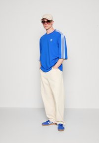 Királykék, oversized T-shirt fehér csíkokkal az ujjakon, bézs sapka és napszemüveg; a krém színű, bő szárú nadrág és a kék sportcipő teszi teljessé a megjelenést.