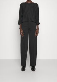 Haut noir ample avec un large col, associé à un pantalon taille haute à jambes droites ; les deux sont en textile lisse et taillé. Des chaussures noires complètent le look.