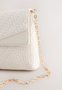 White woven leather handbag with a triangular flap and gold chain strap against a neutral background.