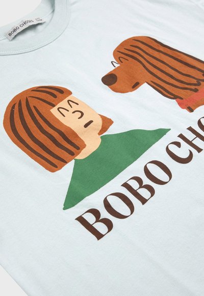 Ilustracja osoby z brązowymi pasmami we włosach i zieloną koszulą, która stoi naprzeciw brązowo pręgowanego psa, poniżej znajduje się tekst "BOBO CHOSES".