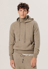 Beige Pullover mit hohem Kragen und befestigter Kapuze. Mit Kordelzügen mit Metallspitzen, gerippten Bündchen und lässiger Passform. Schlichte Textur.