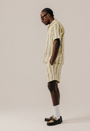Homme aux cheveux tressés portant des lunettes, une chemise jaune à rayures manches courtes et un short assorti, des chaussettes blanches et des mocassins bicolores, debout de profil.