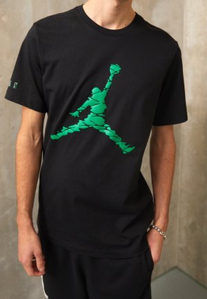 Black t-shirt with green fragmented silhouette of a jumping basketball player on the chest worn by a person.