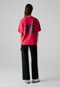Red Bull Racing rosa bomull t-shirt med svart grafisk tryck på baksidan. Modellen har kombinerat den med svarta vida byxor och beige skor.