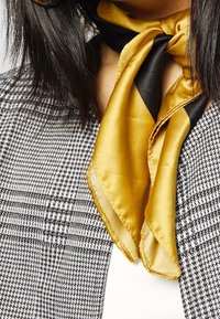 Écharpe en soie aux tons or et noir, nouée autour du cou. En arrière-plan, un blazer à motif pied-de-poule noir et blanc.