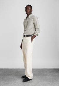 Stripet skjorte med knapper i lysegrå og mørkegrå, kombinert med høyhårede kremfargede bukser og svarte loafers, som gir en strømlinjeformet silhuett.