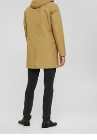 Beżowa, pikowana parka z kapturem o prostym kroju, charakteryzująca się gładką fakturą. Noszona na ciemnych, dopasowanych jeansach i czarnych sneakersach.