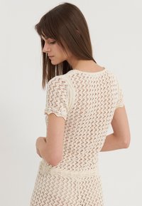 Sweter w kolorze kremowym z teksturowanym wzorem w zygzak, krótkie rękawy i okrągły dekolt. Posiada falbaniaste wykończenia oraz dopasowany krój.