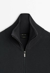 Pull noir en maille côtelée à fermeture éclair avec col montant, partiellement ouvert, montrant l'étiquette de la marque à l'intérieur du col.