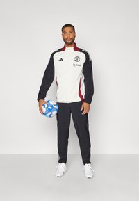 Zwart-witte sportjack met bordeauxrode accenten, adidas-strepen, logo en tekst. Zwarte broek en witte voetbalschoenen. Houdt een blauwe voetbal vast.