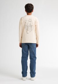 Beige Langarmshirt mit grafischem Druck auf dem Rücken, kombiniert mit blauen Jeans und weißen Sneakers. Einfaches, lässiges Design.