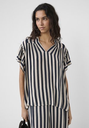 Žena s dlhými tmavými vlasmi, oblečená v voľnom námornícko-modrom a béžovom pruhovanom tričku a zodpovedajúcich nohaviciach, držiaca tmavú tašku.