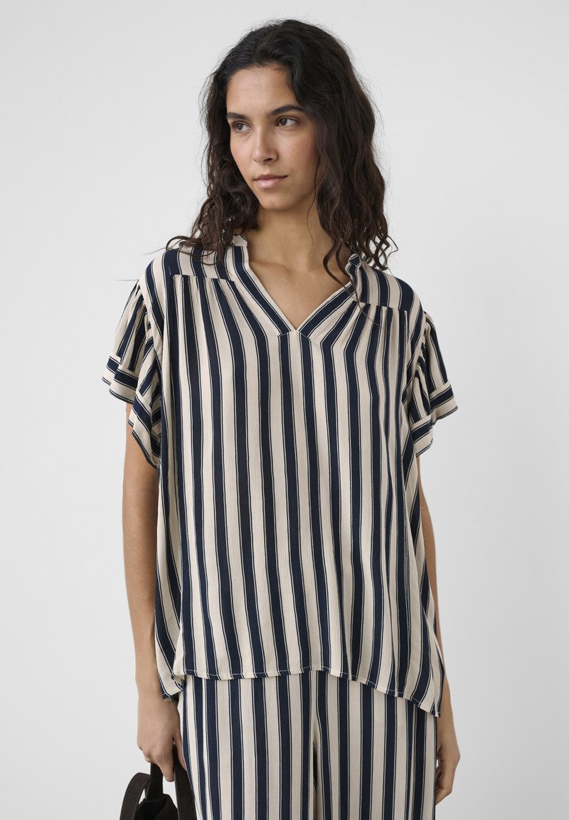 Femme aux cheveux longs et foncés portant un haut ample à rayures verticales bleu marine et beige assorti à un pantalon, tenant un sac sombre.