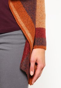 Pletený kardigan s pruhy v oranžové, vínové a námořnické modři. Má měkkou texturu, otevřený přední design a splývavý lem.