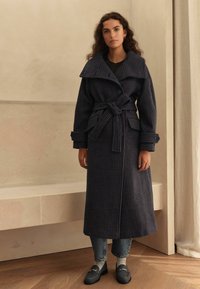 Navy plaid coat with wide collar, belted waist, large front pockets, textured fabric, paired with gray jeans and shoes, standing on wooden floor.
