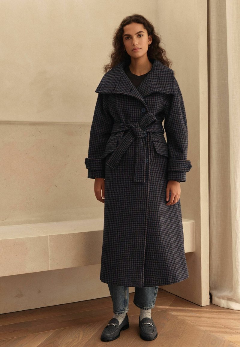 Navy plaid coat with wide collar, belted waist, large front pockets, textured fabric, paired with gray jeans and shoes, standing on wooden floor.