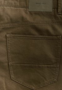 Olíva zöld farmer anyag, kiemelkedő hátsó zsebbel és varrott részletekkel. A zseb felett látható egy "DENIM SUPPLY" felirattal ellátott címke.