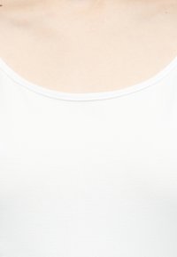 Gros plan sur l'encolure d'un débardeur blanc contre une peau claire, montrant la partie supérieure de la poitrine et la zone des clavicules.