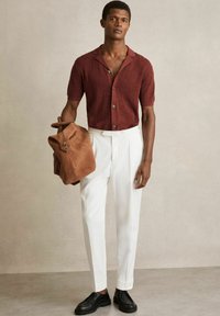 Cămașă tricotată burgundy cu mânecă scurtă și motive hexagonale, asortată cu pantaloni albi evazați și pantofi din piele neagră, ținând un rucsac din suede maro.