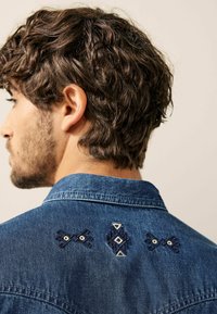 Camisa de mezclilla azul oscuro con cuello puntiagudo y bordados decorativos en la parte posterior con patrones de triángulos y formas geométricas.