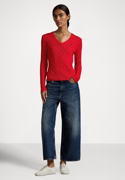 Piros kötött pulóver V-nyakkal, széles szárú sötétkék farmerrel és világos színű sportcipővel párosítva. Egyszerű kialakítás és texturált anyag.
