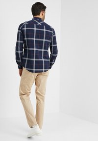 Camicia a quadri blu navy con linee bianche e rosse, maniche lunghe, colletto button-down, abbinata a pantaloni beige e sneakers bianche.