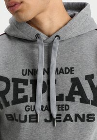 Felpa grigia in cotone, con tasca a marsupio, spessi cordoncini e testo nero audace "REPLAY" e "BLUE JEANS" sul davanti.