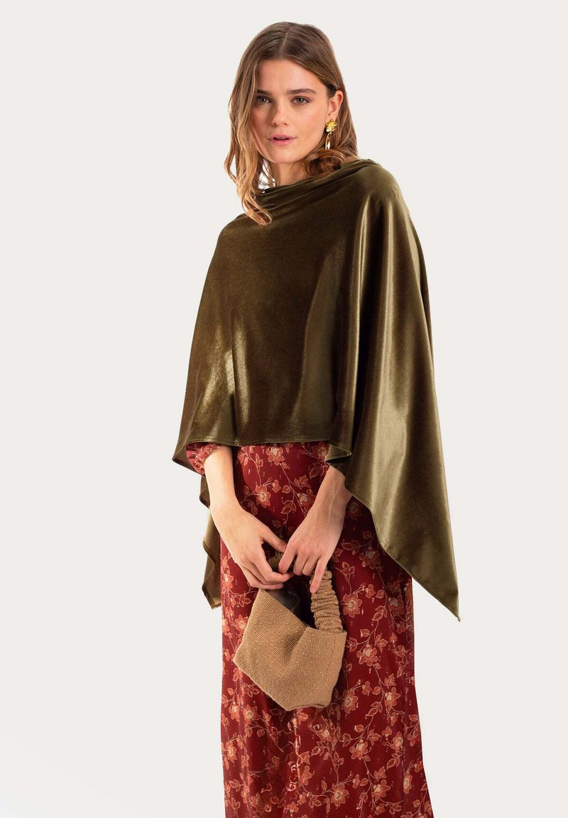 Poncho de terciopelo verde oliva que cae sobre un vestido rojo con estampado floral, con un corte relajado y textura suave. La modelo sostiene un pequeño bolso marrón.