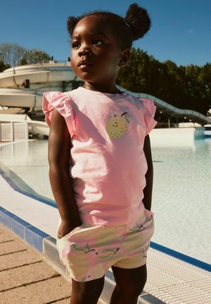Junges Kind mit Zöpfen steht an einem Pool, trägt ein rosa Shirt mit Fruchtmotiv und passende gemusterte Shorts an einem sonnigen Tag.