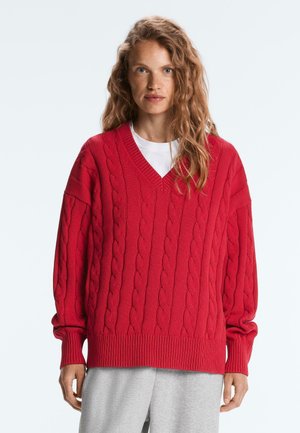 Kvinde med langt krøllet hår iført en stor rød kabelstrikket V-hals sweater og lysegrå bukser, stående mod en ensfarvet hvid baggrund.