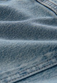 Detailní záběr na světle modrou denimovou látku s viditelným šitím a texturovaným tkaním, ukazující sklady a vrásky.