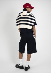 Personne portant un short noir, une chemise noire, des mocassins noirs avec des chaussettes blanches, une casquette rouge et un pull rayé drapé sur les épaules, vue de profil.