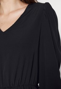 Mustapaita, jossa on V-pääntie, pitkät hihat ja rypytetty vyötärö. Valmistettu sileästä kankaasta, jossa on pehmeä laskeuma ja hienovaraiset olkapään rypytykset.