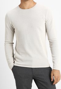 Světlé béžové tričko s dlouhým rukávem s texturovaným vzorem včelích pláství a kulatým výstřihem; přiléhavý střih, zkombinováno s tmavými kalhotami.