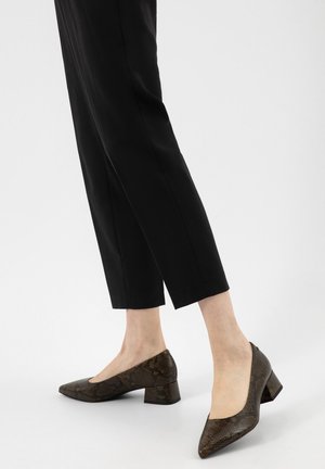 Osoba ubrana w czarne spodnie o długości do połowy łydki oraz brązowe buty na niskim, szerokim obcasie ze wzorem wężowej skóry, stojąca na białej podłodze.