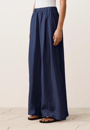 Žena v tmavomodrých širokých nohaviciach s elastickým pásom a čiernych žabkách stojí na svetlej dlažbe.