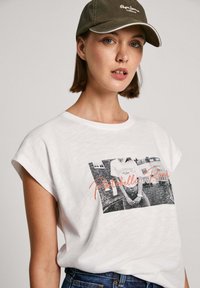 Camiseta de algodón blanca con mangas enrolladas, que presenta una impresión fotográfica en blanco y negro y "Portobello Road" en texto naranja. Ajuste casual y relajado.
