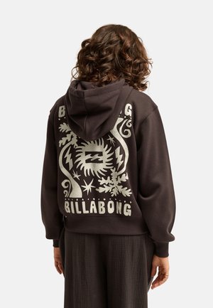 Osoba o kręconych włosach, ubrana w ciemną bluzę z kapturem z graficznymi wzorami i tekstem "Billabong" na plecach, stoi na jednolitym tle.
