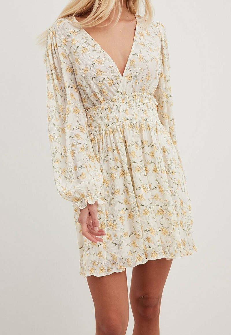 Femme portant une robe crème à fleurs avec de longues manches bouffantes, un profond décolleté en V, une taille smockée et un ourlet court à volants.