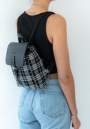 Mochila de cuadros en blanco y negro con una solapa de cuero negro, correas ajustables y un bolsillo con cremallera en la parte frontal. Diseño casual.