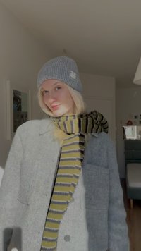 Grå strikket hue, lang stribet halstørklæde i gult og gråt, grå jakke med knapper, blød tekstur, indendørs omgivelser med neutrale toner.