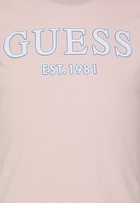 Růžové bavlněné tričko světle růžové barvy s velkým logem "GUESS" v bílé barvě s modrým obrysem a menším písmem "EST. 1981" pod ním. Hladká textura.
