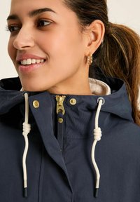 Jūras spēku kapuce uzvalks ar zelta rāvējslēdzēju, baltu šnīri detaļām un mīkstu iekšējo odere. Tuvplāns uz apkakles un sejas.