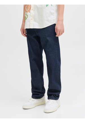 Personne portant un jean bleu foncé ample, des baskets blanches et une chemise blanche à boutons avec un motif floral.