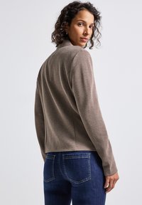Veste grise texturée avec un col, design ajusté et manches longues, associée à un jean bleu foncé. Le tissu présente un motif côtelé.