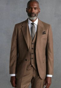 Brown wool suit with a notch lapel, single-breasted jacket, and matching trousers. White shirt and patterned tie. Pocket square in jacket.