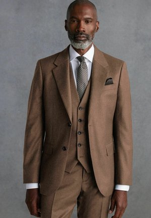 Vestito in lana marrone con rever a punta, giacca monopetto e pantaloni coordinati. Camicia bianca e cravatta a fantasia. Fazzoletto da taschino nella giacca.